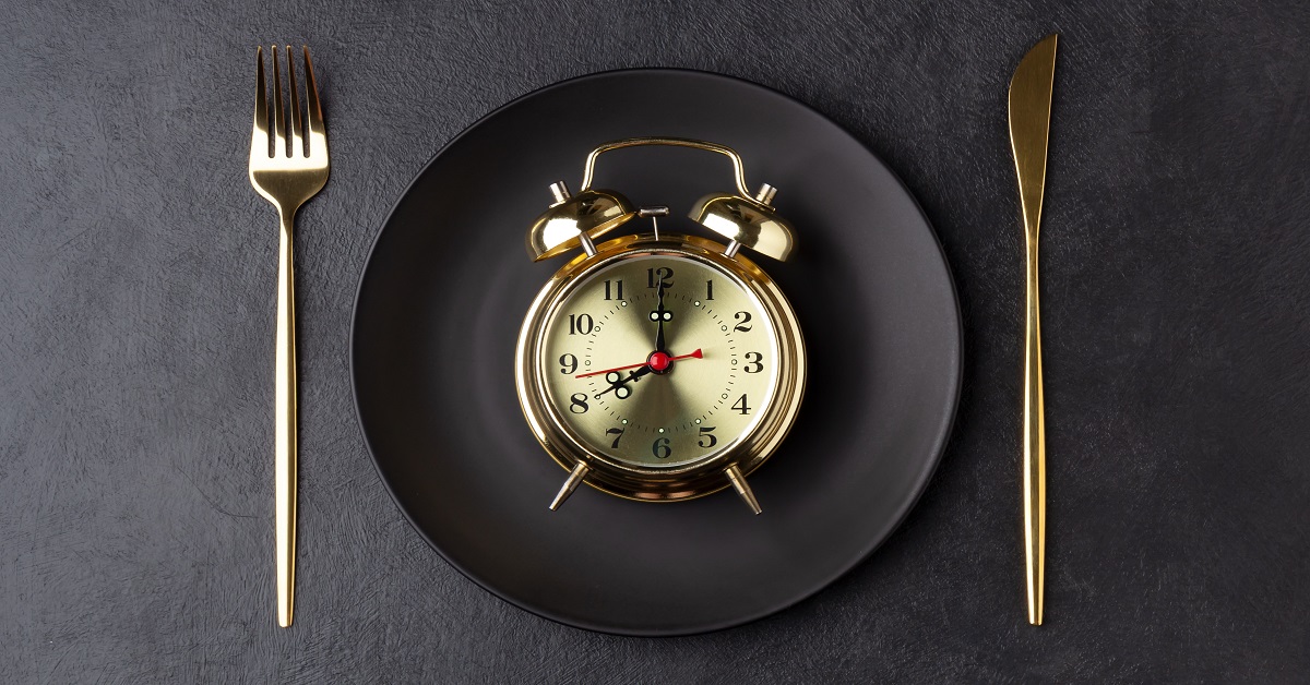 Golden alarm clock on a black plate with a golden knife and fork. Intermittent fasting concept. Horizontal orientation, top view.